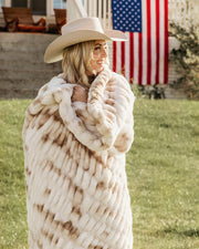 A woman outdoors wrapped in a chunky cream faux-fur throw blanket with tan patches, wearing a light cowboy hat.
