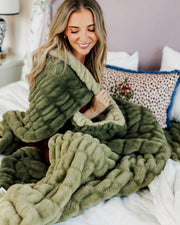 A woman sitting on a bed wrapped in a chunky, ribbed plush throw blanket. The blanket is olive green with an ombre effect.