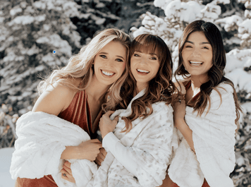 Three smiling women in cozy Minky Couture blankets enjoying a snowy winter day, perfect for wedding gifts.