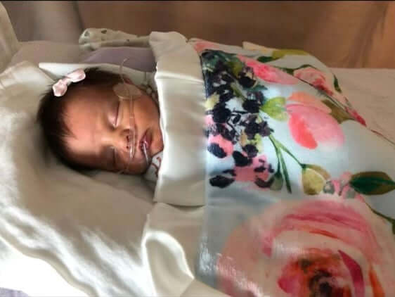Heart of Minky: Evie, a baby in a floral blanket, resting in a hospital setting with medical equipment.