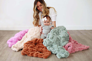 When Can Babies Sleep With a Blanket? Mother and baby surrounded by colorful, soft blankets on the floor.