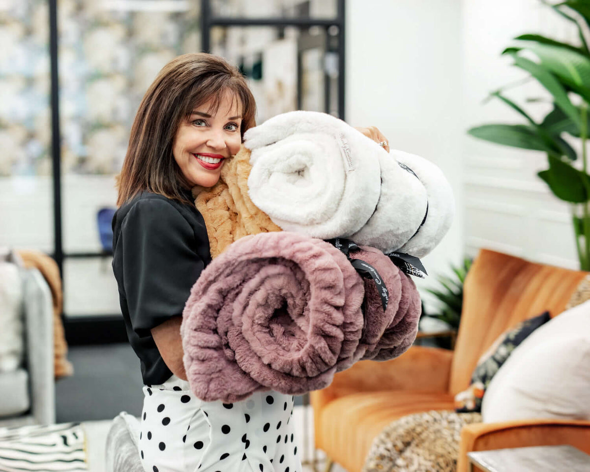 How Did Minky Couture Get Its Start? A woman joyfully holding multiple Minky Couture blankets in a stylish setting.