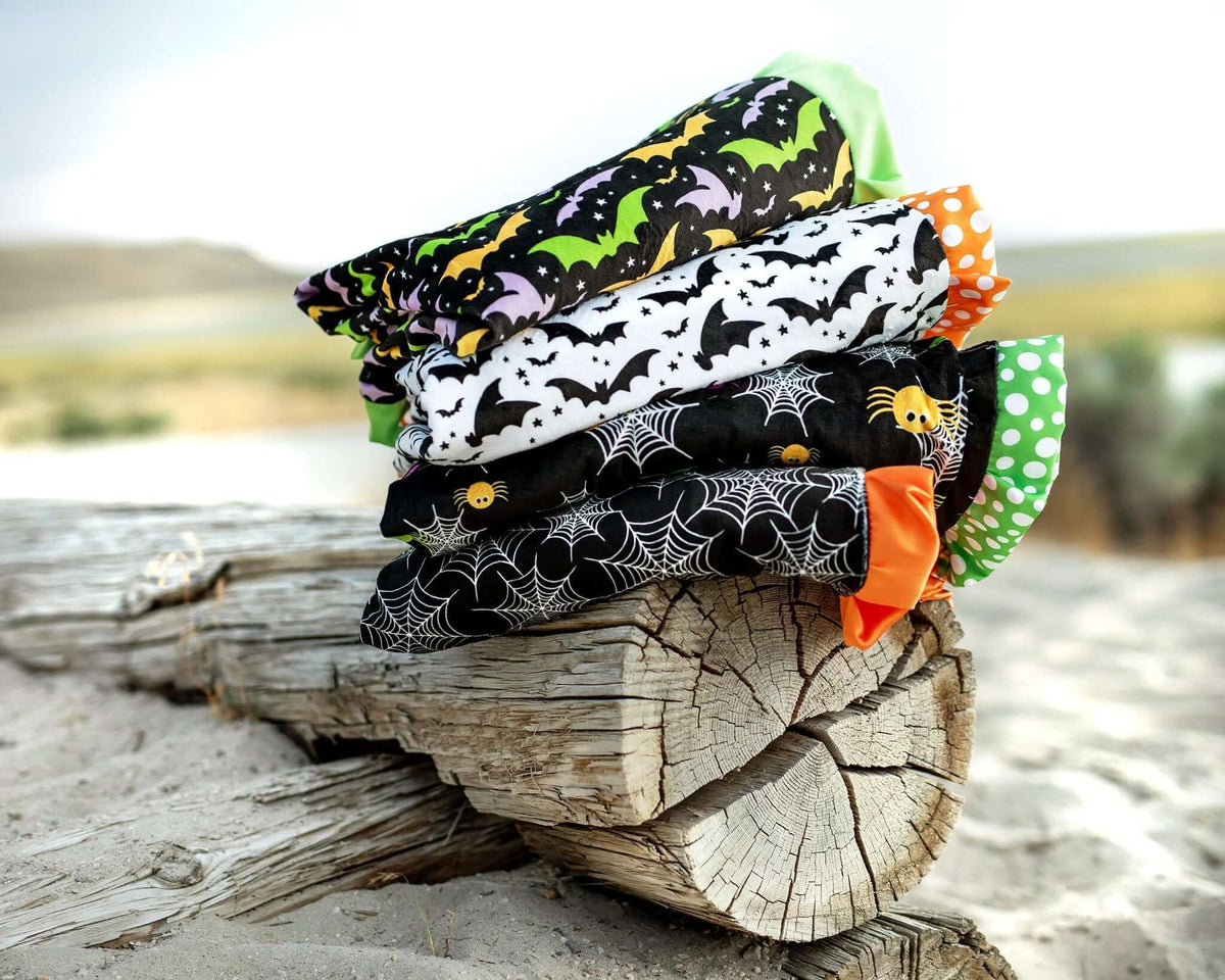 Colorful Halloween blankets in bat and spider patterns stacked on a log outdoors.