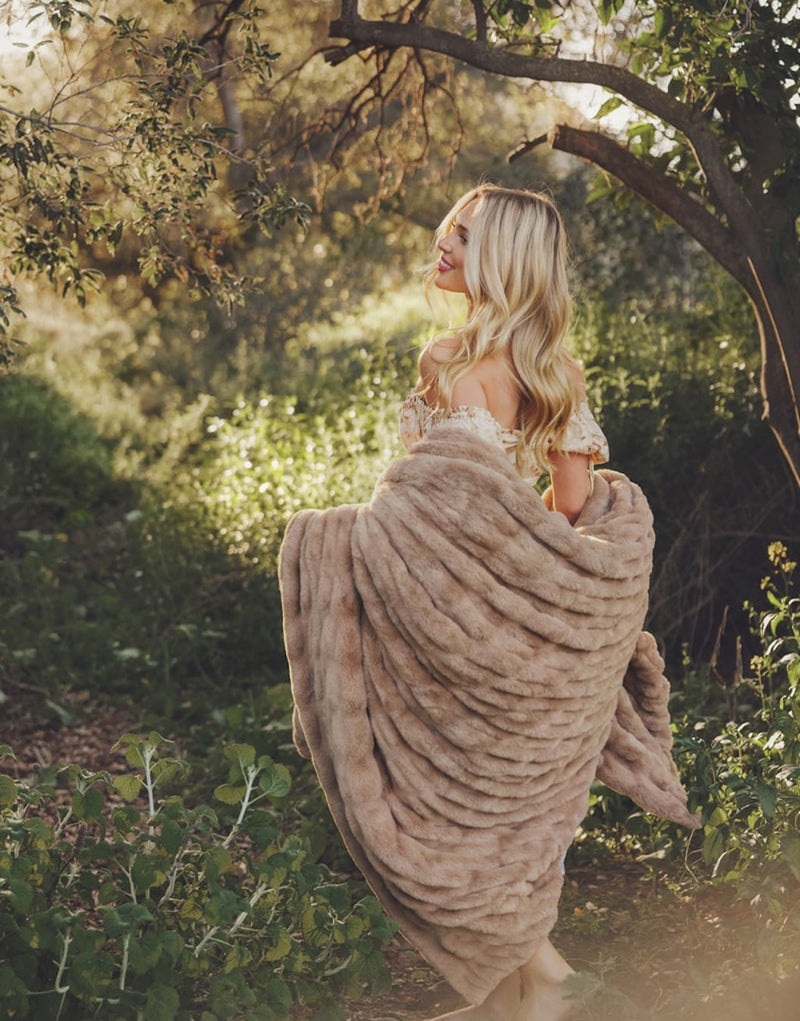 Woman in a flowing dress wrapped in a plush blanket, surrounded by lush greenery, smiling and enjoying nature.