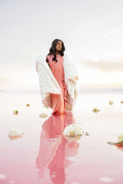 Woman wearing a pink dress and Hugs Cream Blanket walking on pink water, surrounded by flowers.