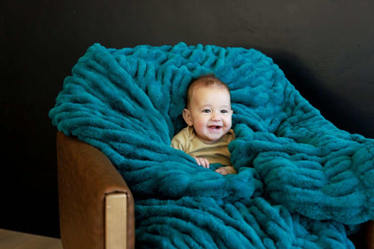 Happy baby snuggled in a Hugs Deep Lake Blanket on a cozy chair, radiating comfort and warmth.