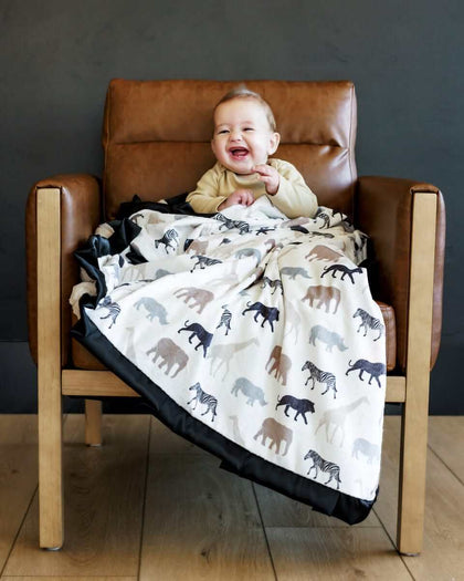 Baby smiling on a chair, wrapped in Safari Blanket featuring animal silhouettes in neutral tones.