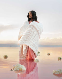 A model wrapped in the Biggest Hugs Cream Blanket stands gracefully in a serene landscape, showcasing comfort and elegance.