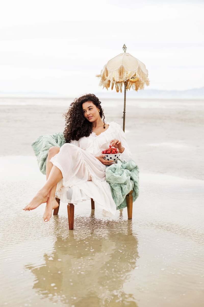 Woman sitting on a beach chair with an umbrella, holding flowers, wearing a white dress and green blanket.