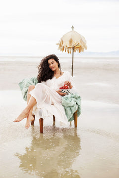 Woman sitting on a beach chair with an umbrella, holding flowers, wearing a white dress and green blanket.