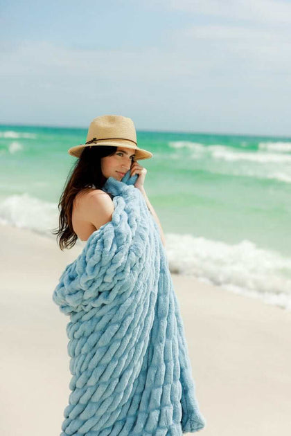 Hugs Alaskan Blue Blanket draped over a woman on the beach, showcasing its plush texture and soothing blue hue.