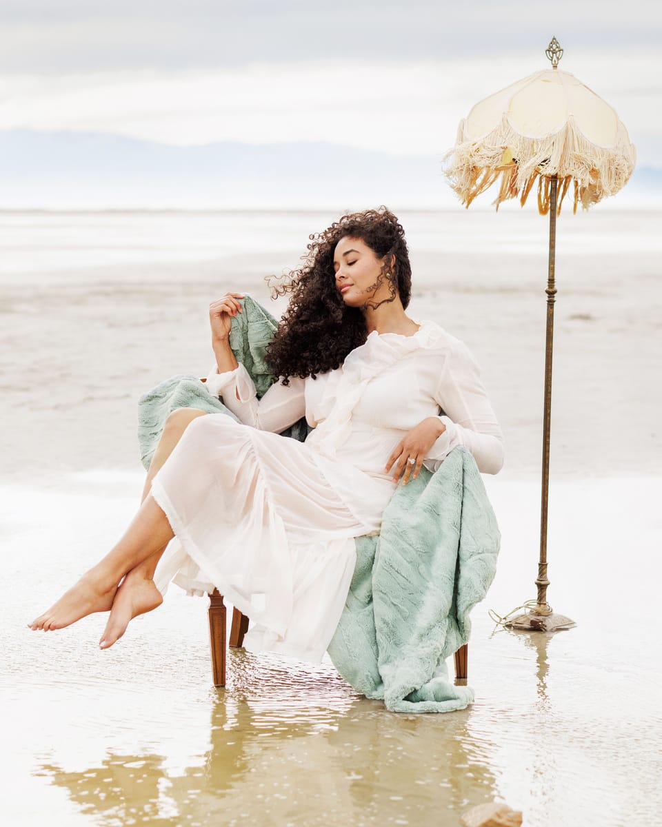 Woman in a white dress sitting on a chair in shallow water with an umbrella in the background on a green soft blanket.