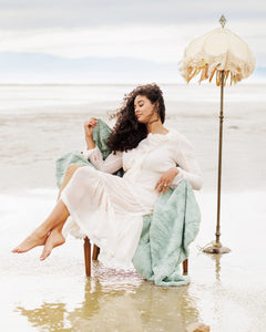 Woman in a white dress sitting on a chair in shallow water with an umbrella in the background on a green soft blanket.