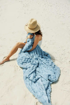 Woman relaxing on the beach wrapped in the Hugs Alaskan Blue Blanket, enjoying sunny vibes.