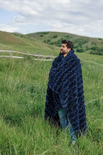 A man wrapped in an Ultra Sorbet Navy Blanket stands in a grassy field, enjoying the cozy warmth of the weighted blanket.