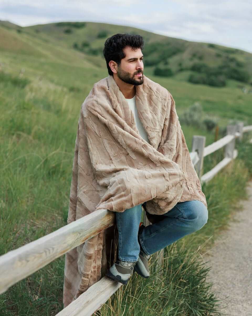 Man sitting on a fence outdoors, wrapped in a Sorbet Milk Chocolate Blanket, enjoying the cozy comfort.