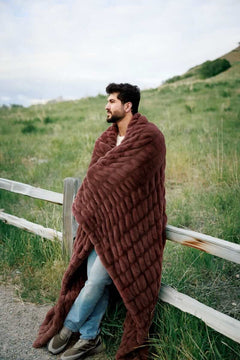 Man wrapped in a warm brown mahogony blanket standing in a grassy field with a white fence.