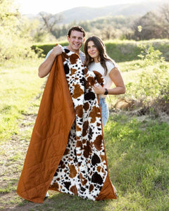 Couple outdoors holding the Everywhere Blanket Brown Cow, featuring cozy cow print and solid camel brown colors.
