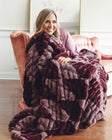 Woman smiling on a sofa, wrapped in a plush Minky Couture Hugs Shelby Mulberry Diamond blanket.