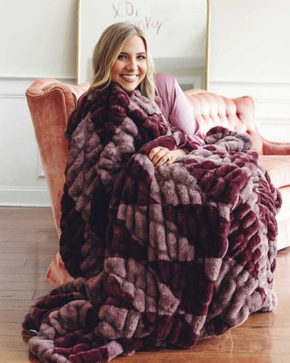 Woman smiling on a sofa, wrapped in a plush Minky Couture Hugs Shelby Mulberry Diamond blanket.
