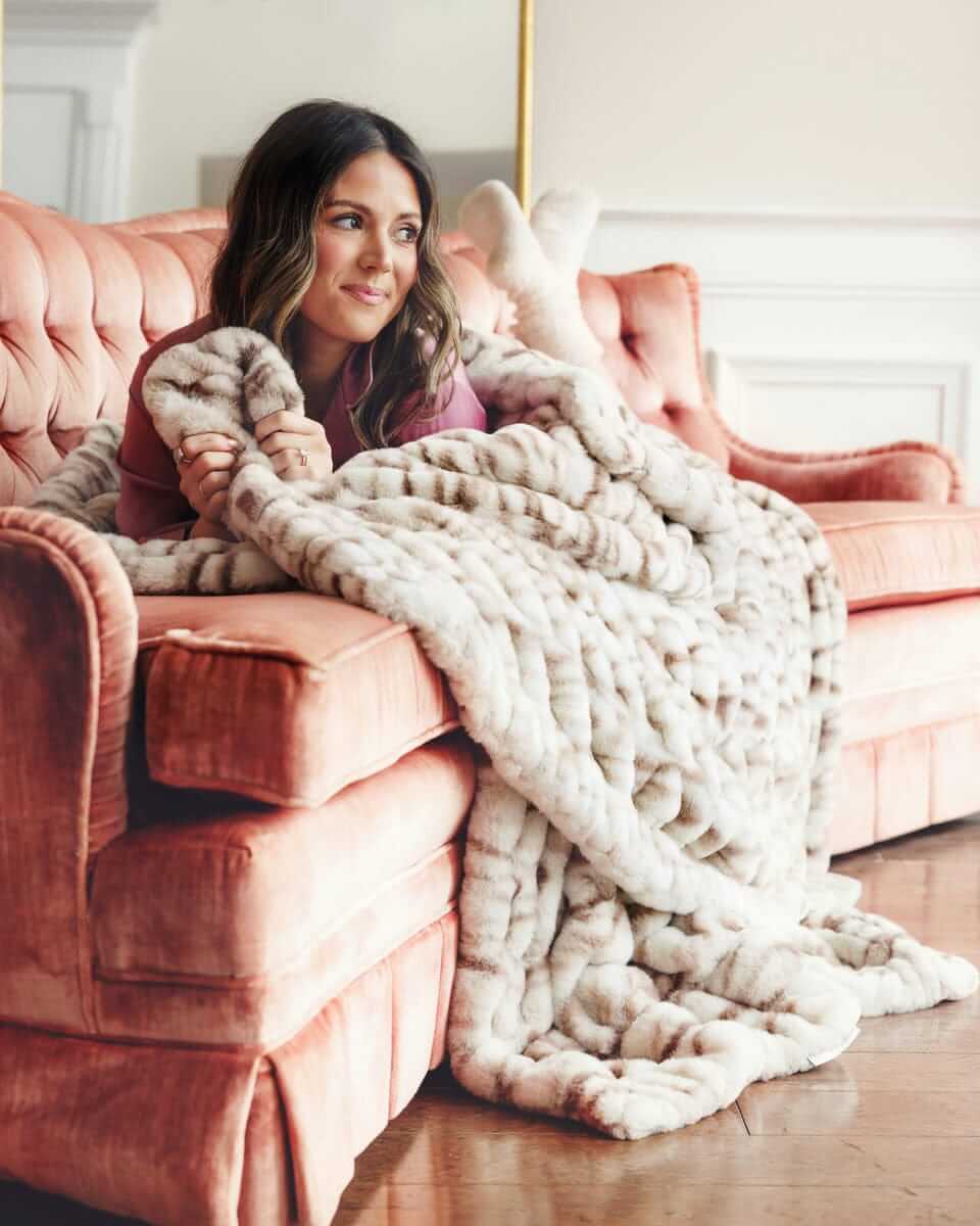Woman lounging on a sofa, wrapped in a faux fur Hugs Kaci Chocolate Marble blanket.