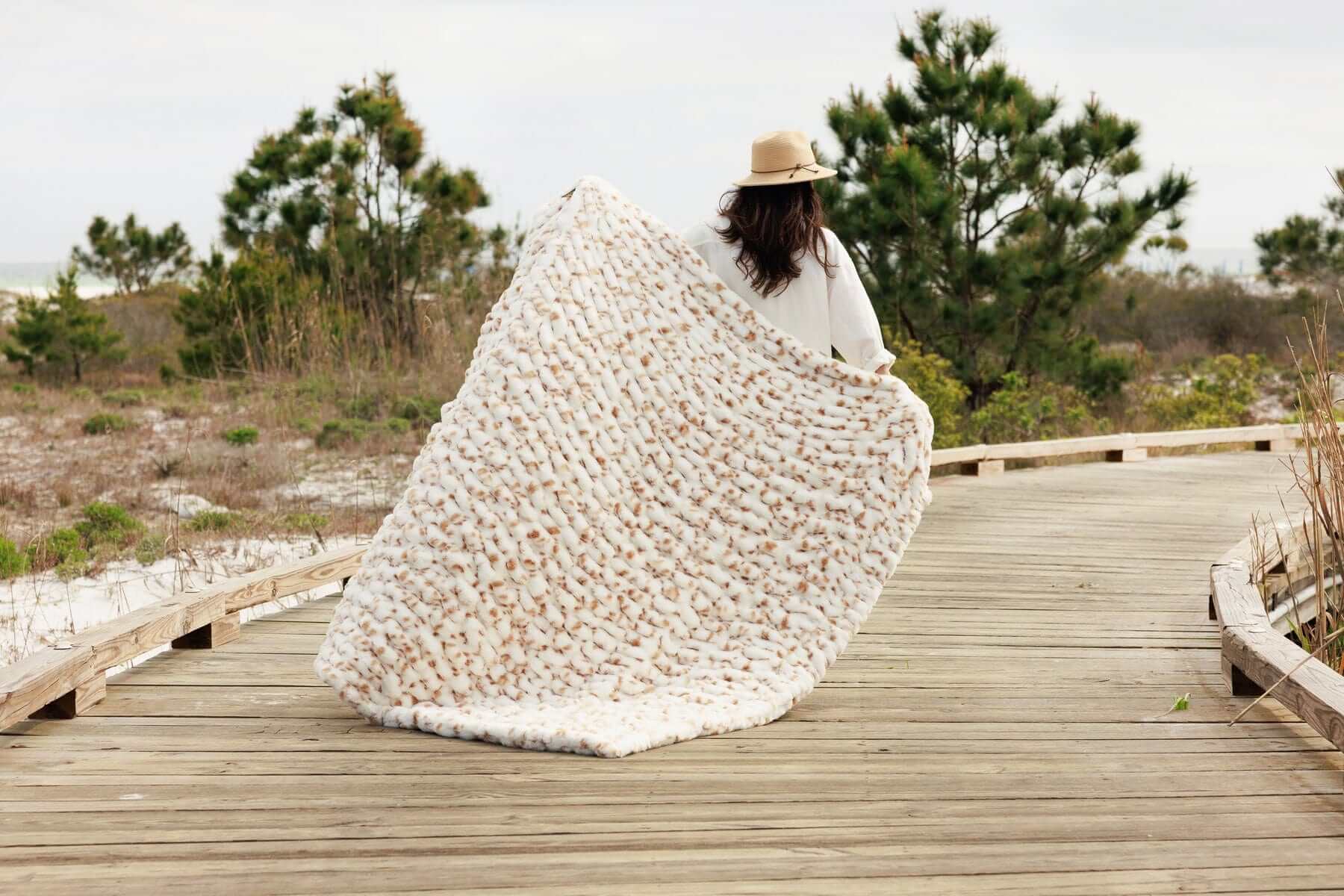 Woman holding the Hugs Spotted Fawn Blanket while walking on a wooden path surrounded by nature.