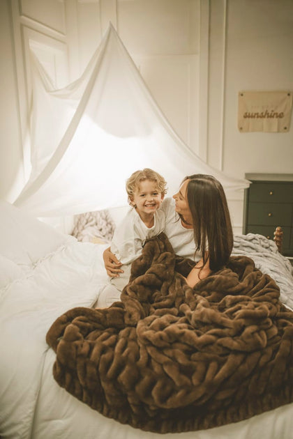 Woman and child sitting on a bed with a brown Hugs Hot Fudge Blanket, smiling.