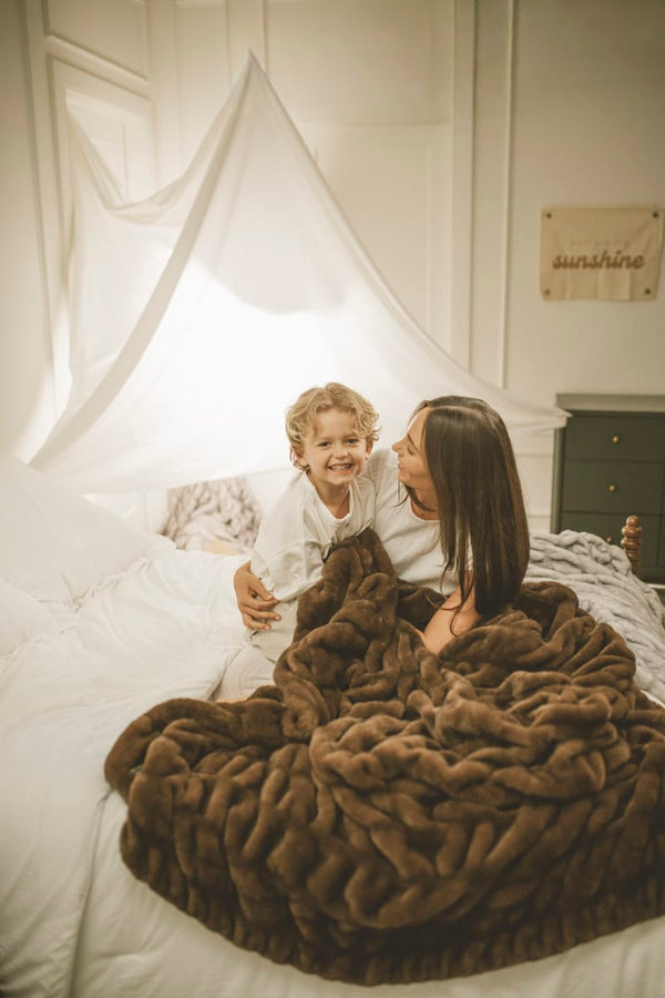 Woman and child sitting on a bed with a brown Hugs Hot Fudge Blanket, smiling.
