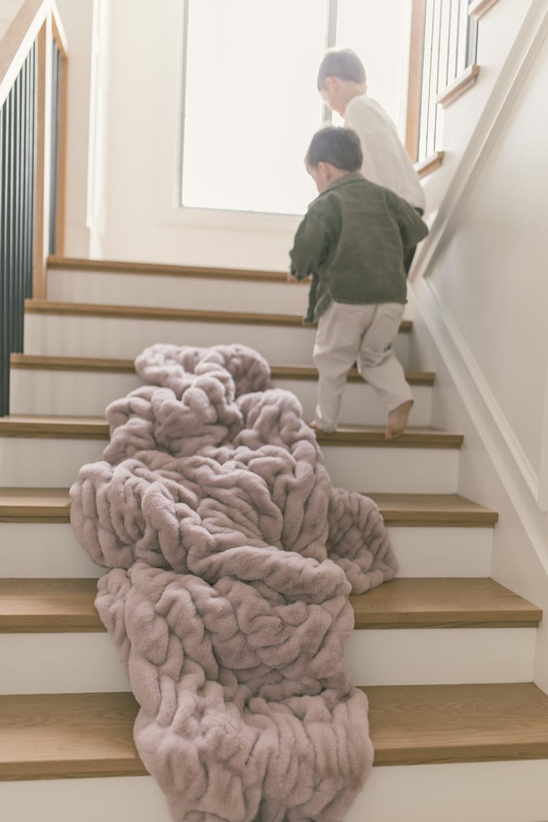 Two children walking up stairs near a soft, plush Hugs Heirloom Rose Blanket draped on the steps.