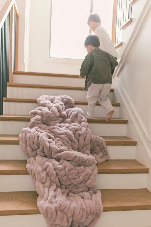 Two children walking up stairs near a soft, plush Hugs Heirloom Rose Blanket draped on the steps.