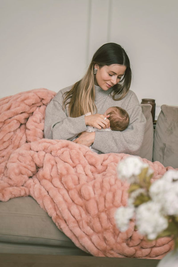 Woman holding a baby on a couch with a pink Hugs Blossom Blanket.