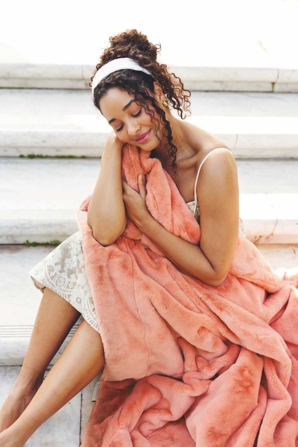 A woman cozying up with the Lush Tawny Spice Blanket in a beautiful coral hue, showcasing its plush texture.