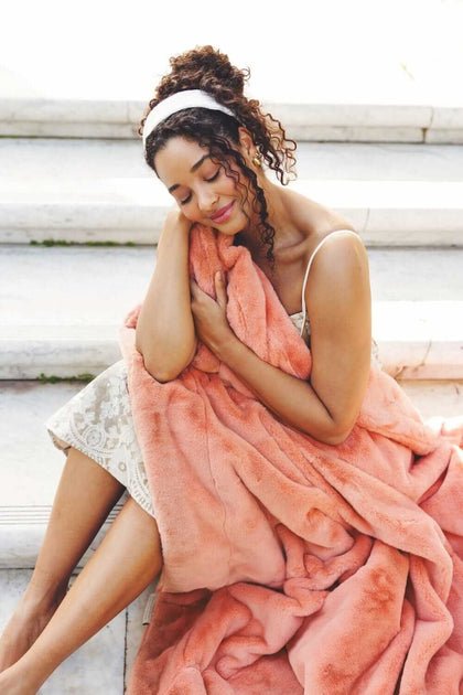 A woman cozying up with the Lush Tawny Spice Blanket in a beautiful coral hue, showcasing its plush texture.