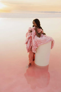 Model wrapped in Hugs Cotton Candy Pink Blanket, standing on a pedestal in a serene pink landscape at sunset.