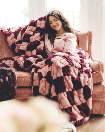 Woman relaxing on a sofa, wrapped in a plush Hugs Devin Pink Checkered blanket.