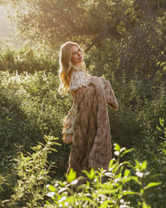 A woman in a sunny outdoor setting holds a Hugs Milk Chocolate Blanket, showcasing its plush texture and warm brown color.