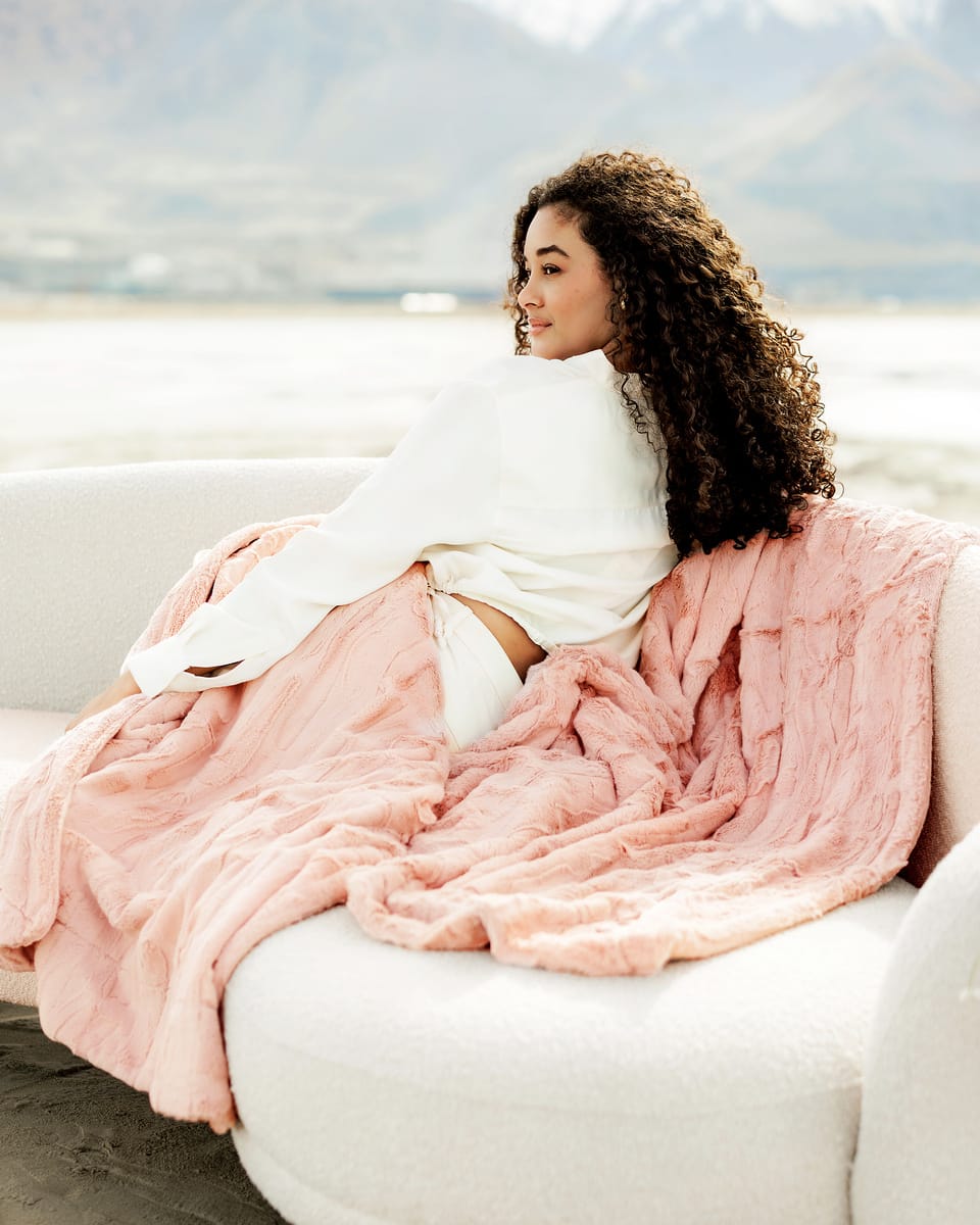 Woman wrapped in a soft Sorbet Apricot Vanilla Blanket on a cozy couch in a scenic outdoor setting.