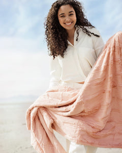A smiling woman holding the Sorbet Apricot Vanilla Blanket with a soft light pink hue, perfect for cozying up.