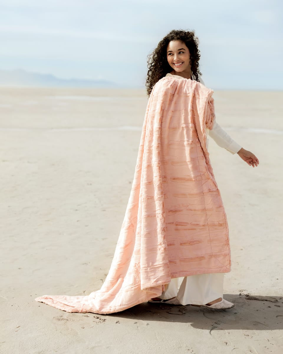 A person wearing the Sorbet Apricot Vanilla Blanket, enjoying a serene beach view, exuding warmth and comfort.