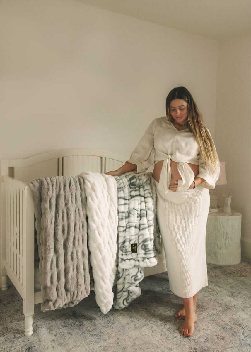 Woman standing beside a crib draped with soft plush Minky Couture blankets in neutral tones inside a calm, minimalist nursery.