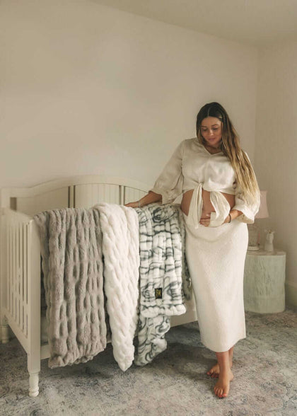 Woman standing beside a crib draped with soft plush Minky Couture blankets in neutral tones inside a calm, minimalist nursery.