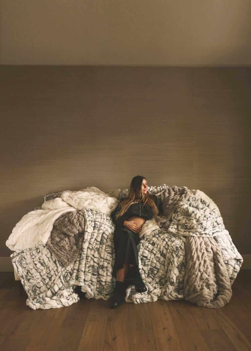 Woman seated on a sofa layered with oversized faux fur Minky Couture blankets in soft neutral tones, styled in a minimalist living space.