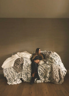 Woman seated on a sofa layered with oversized faux fur Minky Couture blankets in soft neutral tones, styled in a minimalist living space.