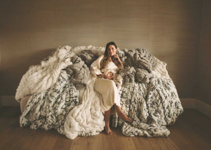 Woman seated on a sofa surrounded by oversized faux fur Minky Couture blankets in a warm, cozy living room setting.
