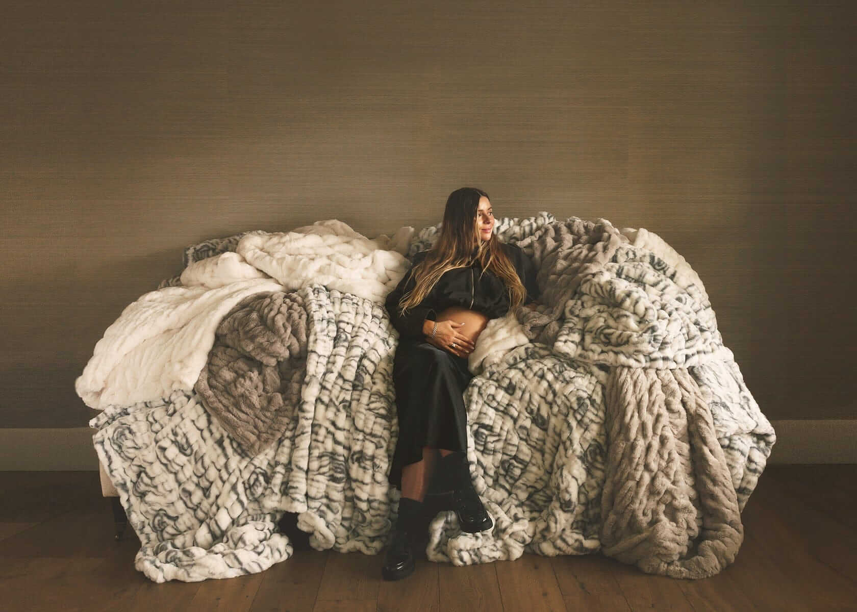 Woman sitting on a couch surrounded by layered faux fur Minky Couture blankets in neutral tones, creating a cozy, relaxed home setting.