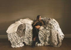 Woman sitting on a couch surrounded by layered faux fur Minky Couture blankets in neutral tones, creating a cozy, relaxed home setting.