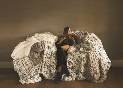 Woman sitting on a couch surrounded by layered faux fur Minky Couture blankets in neutral tones, creating a cozy, relaxed home setting.