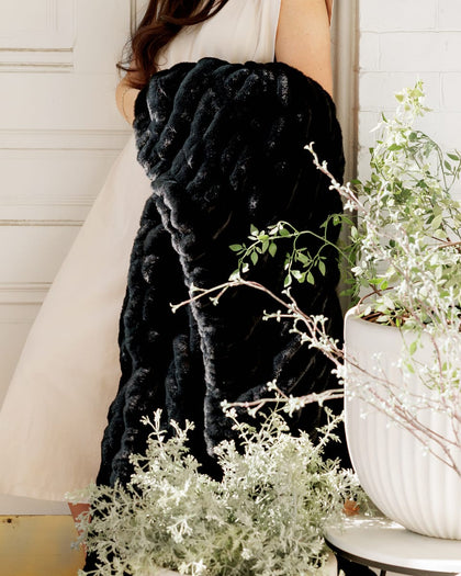 Woman holding the Hugs Black Blanket next to decorative plants in a cozy setting.