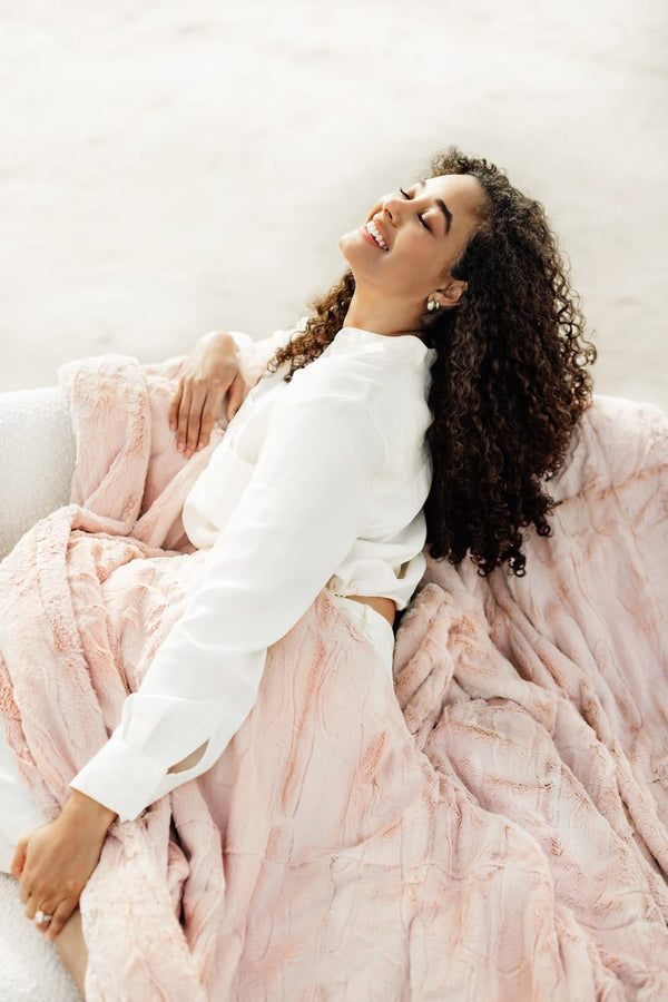 Woman enjoying the warmth of the Sorbet Blush Pink Blanket while relaxing on a cozy sofa.