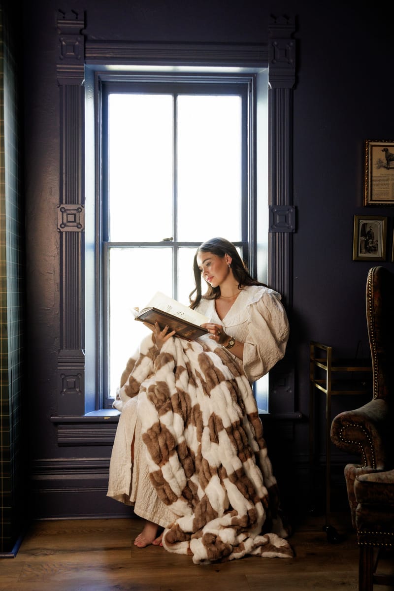 A woman enjoys a book while wrapped in the cozy Hugs Check Milk Chocolate Blanket by a sunny window.
